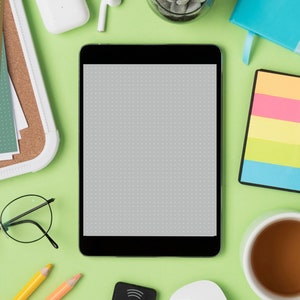 May include: A flat lay image of a workspace with a tablet, a cup of coffee, a notepad, a pair of glasses, pencils, and other office supplies. The tablet is black and has a blank screen. The cup of coffee is white and has a brown liquid inside. The notepad is black and has a colorful sticky note on it. The glasses are black and have a round frame. The pencils are yellow and orange. The office supplies are on a green surface.