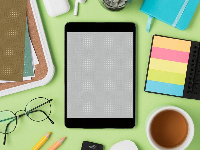 May include: A flat lay image of a workspace with a tablet, a cup of coffee, a notepad, a pair of glasses, pencils, and other office supplies. The tablet is black and has a blank screen. The cup of coffee is white and has a brown liquid inside. The notepad is black and has a colorful sticky note on it. The glasses are black and have a round frame. The pencils are yellow and orange. The office supplies are on a green surface.