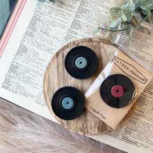 May include: Three black vinyl record lapel pins with colored centers, one red, one light blue, and one light green, arranged on a wooden surface. The pins are packaged with a "Hayden Handcraft" label. The background includes a vintage book and greenery.
