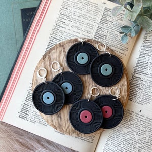 May include: Black vinyl record earrings with silver-colored leverback closures. Each earring features a black disc with a colored center, including blue, red, and green. The earrings are displayed on a wooden surface with an open book.