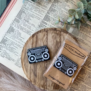 May include: Black and white boombox fridge magnets. One magnet is displayed on a wooden surface, the other is in a clear package with the text "HAYDEN | Handcraft Fridge Magnet". The background includes a vintage book and greenery.