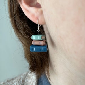 Puede incluir: Pendientes de gancho plateados con una pila de tres libros en miniatura. Los libros son de tonos azul, rosa y verde, con líneas blancas en los lomos. Un accesorio encantador para los amantes de la lectura.