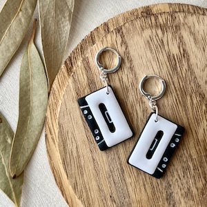 May include: A pair of black and white cassette tape earrings with silver hoops. The earrings are on a wooden surface with dried leaves in the background. The earrings are a unique accessory.