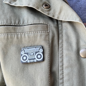 May include: A black and white boombox pin is attached to an olive green jacket. The pin features a detailed outline of a retro stereo with speakers and buttons. The jacket has a zipper, snap buttons, and a pocket.