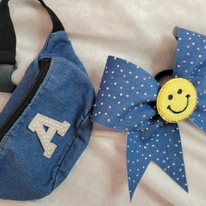 May include: A blue denim fanny pack with a rhinestone letter A and a blue denim hair bow with a yellow smiley face.