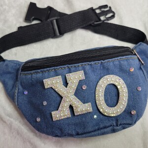 May include: A blue denim fanny pack with a zipper closure and a white pearl embellished "XO" design.