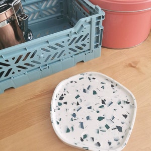 May include: A white, irregular-shaped tray with a speckled pattern of black and green flecks. The tray is made of terrazzo and is perfect for serving snacks or drinks.