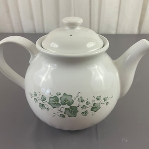 May include: White ceramic teapot with a green ivy design. The teapot has a lid and a spout.