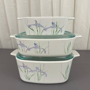 May include: A set of three white ceramic baking dishes with blue and purple iris flower designs. The dishes are stacked on top of each other, with the largest dish on the bottom and the smallest dish on top.