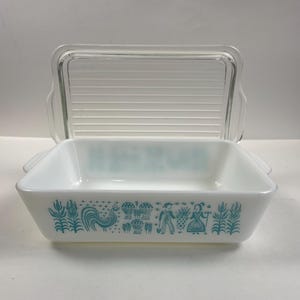 Peut inclure: Un plat de cuisson rectangulaire en verre opalin blanc avec un motif bleu sarcelle représentant des coqs, des épis de blé et des personnages. Le plat comprend un couvercle en verre transparent strié avec des poignées intégrées.