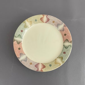 May include: A round, off-white ceramic plate with a decorative border. The border features a repeating pattern of abstract shapes in muted colors including green, pink, and brown. The plate has a smooth, glossy finish.
