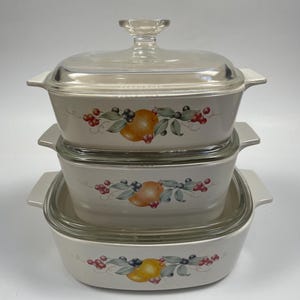 May include: A set of three white ceramic baking dishes with glass lids. The dishes feature a floral design with fruit and berries.