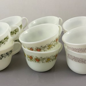 Puede incluir: Un juego de diez tazas de café de cerámica blanca con diseños florales. Las tazas tienen un diseño floral verde y marrón sobre fondo blanco.