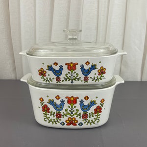May include: A white two-piece casserole dish set with a glass lid. The dish features a colourful floral and bird design with blue birds, red and orange flowers, and green leaves.