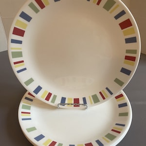 Puede incluir: Una pila de cuatro platos de cerámica blanca con un patrón geométrico colorido alrededor del borde. El patrón presenta rayas rojas, amarillas, azules y verdes.