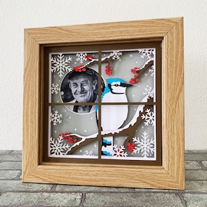 May include: A framed paper craft with a blue jay perched on a branch with red berries. The background is a snowy scene with snowflakes. A man's face is visible in a circle cutout in the upper left corner.