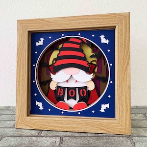 May include: A framed papercraft scene of a gnome dressed as a vampire with a red cape and a black and red striped hat. The gnome is holding a banner that says "BOO" and is standing in front of a full moon with bats flying around it.
