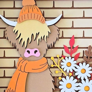 May include: A layered paper art piece featuring a Highland cow wearing an orange hat and scarf. The cow has a light brown body, a pink nose, and white horns. White daisies and fall foliage are in the lower right corner, set against a brick wall background.