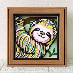 May include: A colourful papercraft sloth hanging in a wooden frame. The sloth is made of layered paper and has a rainbow colour scheme. The frame is brown wood with a black mount.