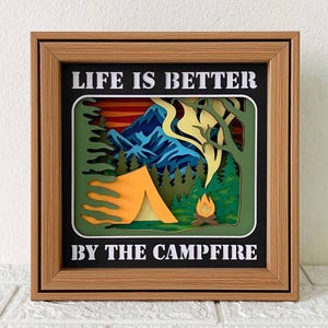 Puede incluir: Una obra de arte en papel en capas enmarcada con un tema de acampada. La obra presenta una tienda de campaña, una fogata, montañas y árboles, con el texto "LIFE IS BETTER BY THE CAMPFIRE" en blanco. El marco es de color marrón claro.