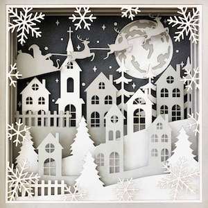 May include: A white papercraft scene depicting a winter village with houses, trees, and a full moon. Santa Claus is flying in his sleigh pulled by reindeer in the sky above the village.