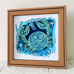 May include: A wooden frame with a blue and green paper cut-out of a crab in a sea-like setting. The crab is intricately designed with swirls and patterns.