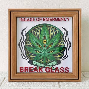 Könnte beinhalten: Gerahmtes Kunstwerk mit einem geschichteten Design einer grünen Cannabisblatt mit gelben Augen. Der Text "IN CASE OF EMERGENCY" und "BREAK GLASS" ist rot. Zwei brennende Joints befinden sich unten, mit Rauch, der auf beiden Seiten aufsteigt.