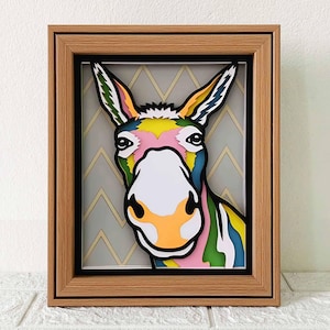 Puede incluir: Un recorte de papel colorido de la cabeza de un burro en un marco de madera. El burro es rosa, amarillo, verde y azul. El fondo es un patrón de chevron gris.