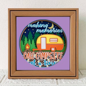 Puede incluir: Obra de arte en papel enmarcada con temática de camping. El diseño presenta una caravana amarilla y naranja, una fogata y el texto "making memories one campsite at a time" sobre un fondo morado, enmarcado en un marco de madera.