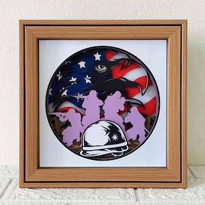 May include: A framed paper art piece with a 3D effect. The artwork features a bald eagle with its wings spread, a US flag, and soldiers in silhouette. The eagle's eye is visible, and the soldiers are holding rifles. The artwork is framed in a light brown wood frame.