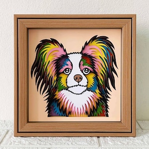 May include: A framed paper art piece of a colorful papillon dog. The dog's fur is layered with different colors, including pink, yellow, green, blue, and black. The dog's eyes are black and white, and its nose is pink. The frame is made of wood and has a light brown finish.