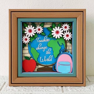 May include: A framed 3D paper craft with a globe, a backpack, an apple, and flowers. The globe is green and blue with the text "Teachers change the world".