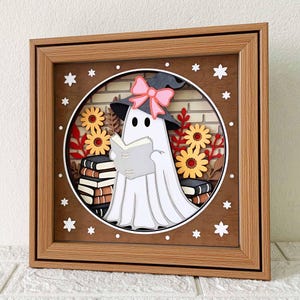 Op de afbeelding: Een witte geest met een roze strik en een heksenhoed leest een boek in een bruin houten frame met een bakstenen achtergrond en herfstbladeren.