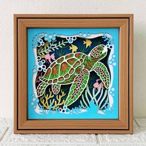 May include: A framed papercraft artwork depicting a green sea turtle swimming in a blue ocean with coral reefs and fish. The turtle has a detailed shell with brown and green markings.
