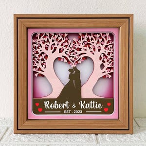 May include: A wooden frame with a pink background and a cutout design of two trees forming a heart shape. The trees are decorated with small pink hearts. Inside the heart, there is a silhouette of a couple. The frame has the text "Robert & Katie EST. 2023" at the bottom.