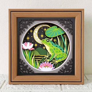 Peut inclure: Une œuvre d'art encadrée en papier représentant une grenouille verte sur une feuille de nénuphar, sur fond de ciel nocturne avec un croissant de lune et des étoiles. L'œuvre comprend des fleurs de lotus roses et du feuillage vert, le tout dans un cadre décoratif.