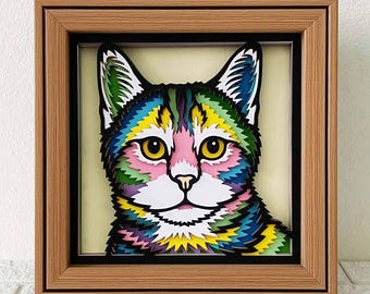 Può includere: Opera d'arte incorniciata con un ritratto di gatto colorato realizzato con carta a strati. Il volto del gatto è raffigurato con vivaci tonalità di rosa, giallo, blu, verde e viola. L'opera è racchiusa in una cornice di legno con un bordo interno nero.