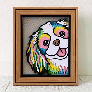 May include: A colorful paper cut-out of a dog's face peeking out from behind a chevron patterned background. The dog's fur is layered in shades of green, yellow, pink, and blue. The dog has a pink tongue sticking out.