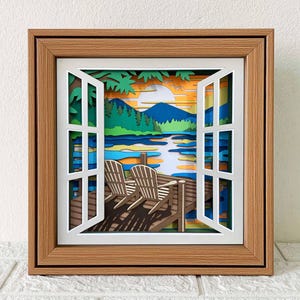 May include: A framed paper art piece depicting a serene lake scene viewed through an open window. The artwork features two chairs on a wooden dock, with a colorful landscape of mountains, trees, and water. The frame is a warm brown color.