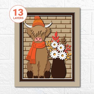 May include: A layered paper art piece featuring a Highland cow wearing an orange hat and scarf, set against a brick wall background. A vase of white daisies and fall foliage is to the right. The artwork is framed in a neutral tone.
