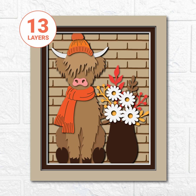May include: A layered paper art piece featuring a Highland cow wearing an orange hat and scarf, set against a brick wall background. A vase of white daisies and fall foliage is to the right. The artwork is framed in a neutral tone.