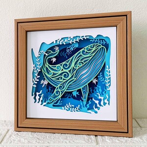 May include: A wooden frame with a white mat and a blue and green paper cut-out of a whale swimming in the ocean. The whale is surrounded by seaweed and fish.