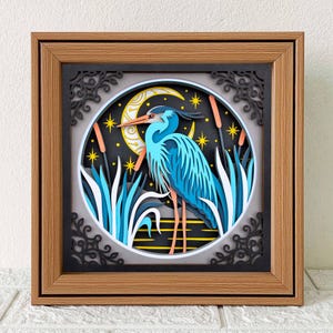 May include: A layered paper art piece featuring a blue heron standing in a marsh under a crescent moon and stars. The artwork is framed in a brown wooden frame with a black decorative border. The heron has orange legs and beak.