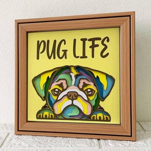 May include: A wooden frame with a yellow background and the text "PUG LIFE" in black. A colorful, layered paper cut-out of a pug dog's face is in the center of the frame.