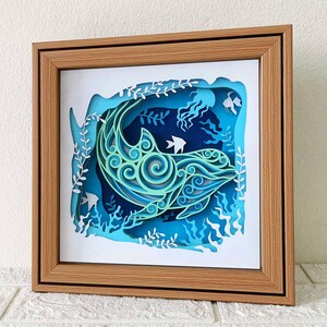 May include: A wooden frame with a blue and green paper cut-out of a dolphin swimming in the ocean. The dolphin is surrounded by seaweed and other sea life.