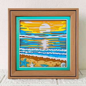 May include: A framed, layered paper art piece depicting a beach sunset. The artwork features a vibrant color palette of yellow, orange, blue, and brown. The scene includes a sun, clouds, ocean waves, and seashells. The frame is a light brown color.