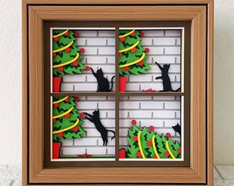 May include: A framed papercraft artwork depicting a window scene with four black cats decorating Christmas trees. The trees are green with red ornaments and yellow lights. The background is a white brick wall.