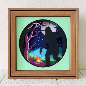 Puede incluir: Una caja de sombra de papel con una silueta de Bigfoot de pie en un bosque por la noche. La escena incluye una fogata con dos personas sentadas junto a ella, una luna llena y estrellas en el cielo.