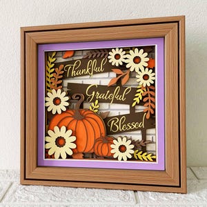 May include: A framed paper craft with a fall theme. The craft features a brick wall background with the words "Thankful", "Grateful", and "Blessed" written on banners. There are also pumpkins, leaves, and flowers in the design.