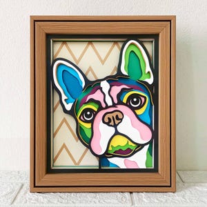 May include: A framed artwork depicting a French Bulldog's face in a layered paper design. The dog is depicted in bright colours including blue, pink, green, and yellow, with a white and black outline. The background features a beige chevron pattern.